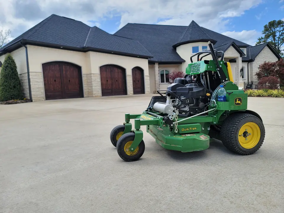 Green stand-on mower on a concrete driveway in front of a large stone house